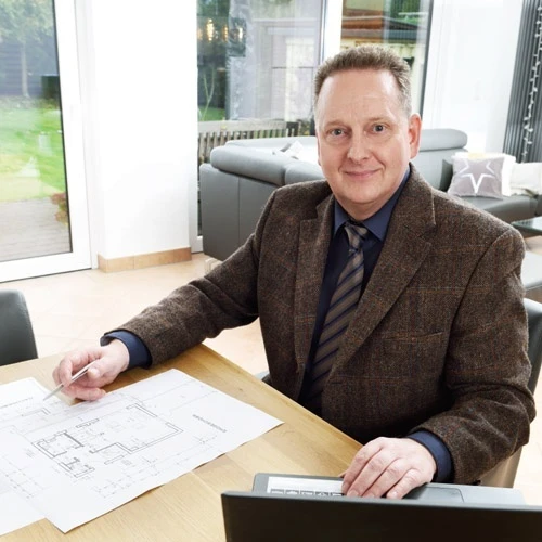 Raimund Klein Immobilien 1 Immobilienmakler sitzt an einem Tisch mit Laptop und Grundrissplan und blickt freundlich in die Kamera.